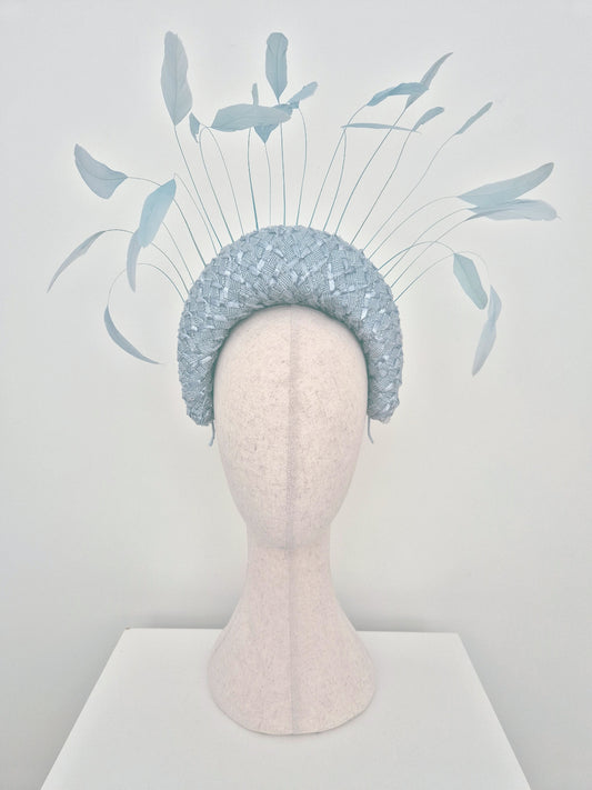 A light blue feathered fascinator halo displayed on a cream coloured mannequin head  against a white studio background.