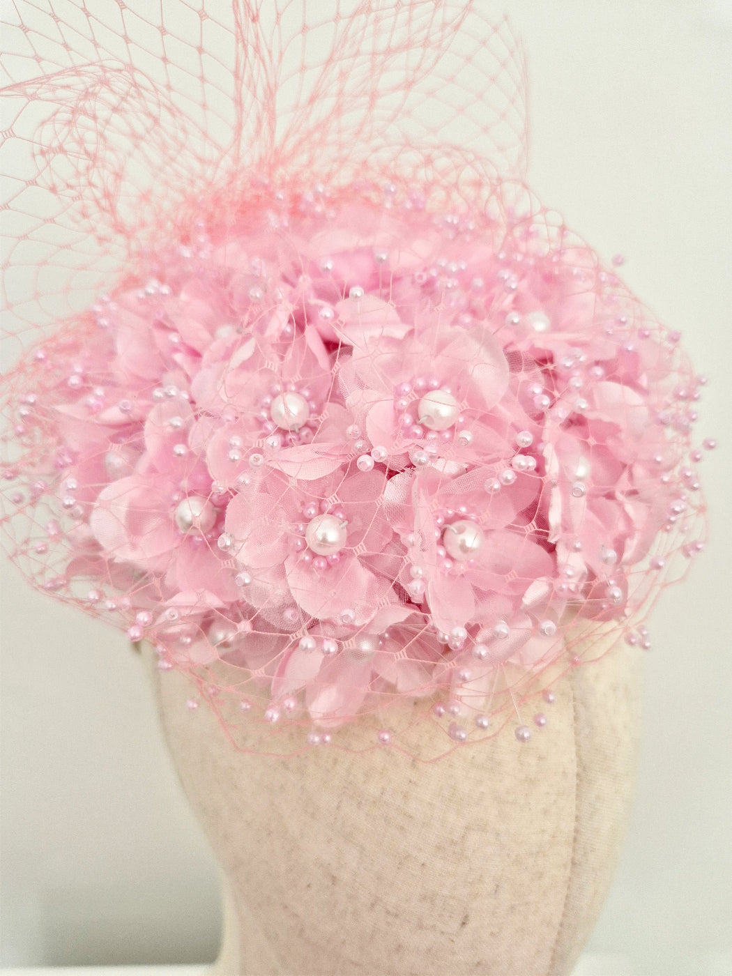 A light pink fascinator hat with layered fabric flowers, pearl-like beads, and a sheer pink netting veil, displayed on a textured cream mannequin head against a plain light background.close up