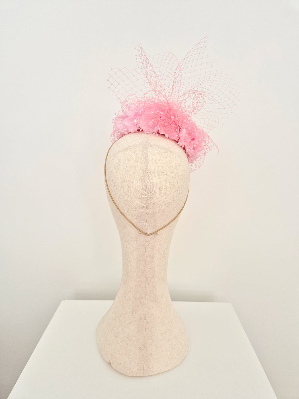 A light pink fascinator hat with a delicate pink mesh veil, displayed on a cream-colored felt mannequin head against a white background.back view