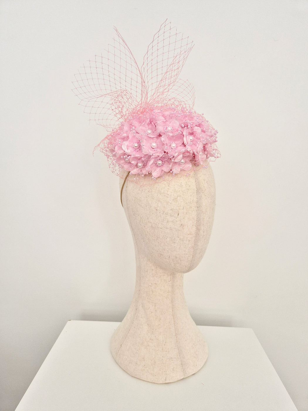A light pink fascinator hat with a floral crown and sheer netting, displayed on a cream-colored felt mannequin head against a plain white background.side view