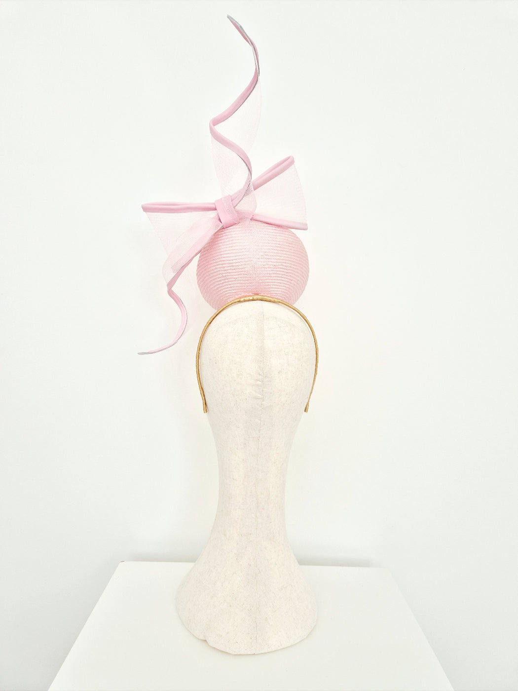 A light pink fascinator headpiece with a large matching pink bow on a mannequin head against a plain white background back view