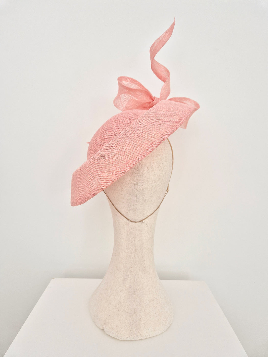 A light pink fascinator hat with a large bow and a single curling feather, displayed on a cream-colored felt mannequin head against a white background. back view