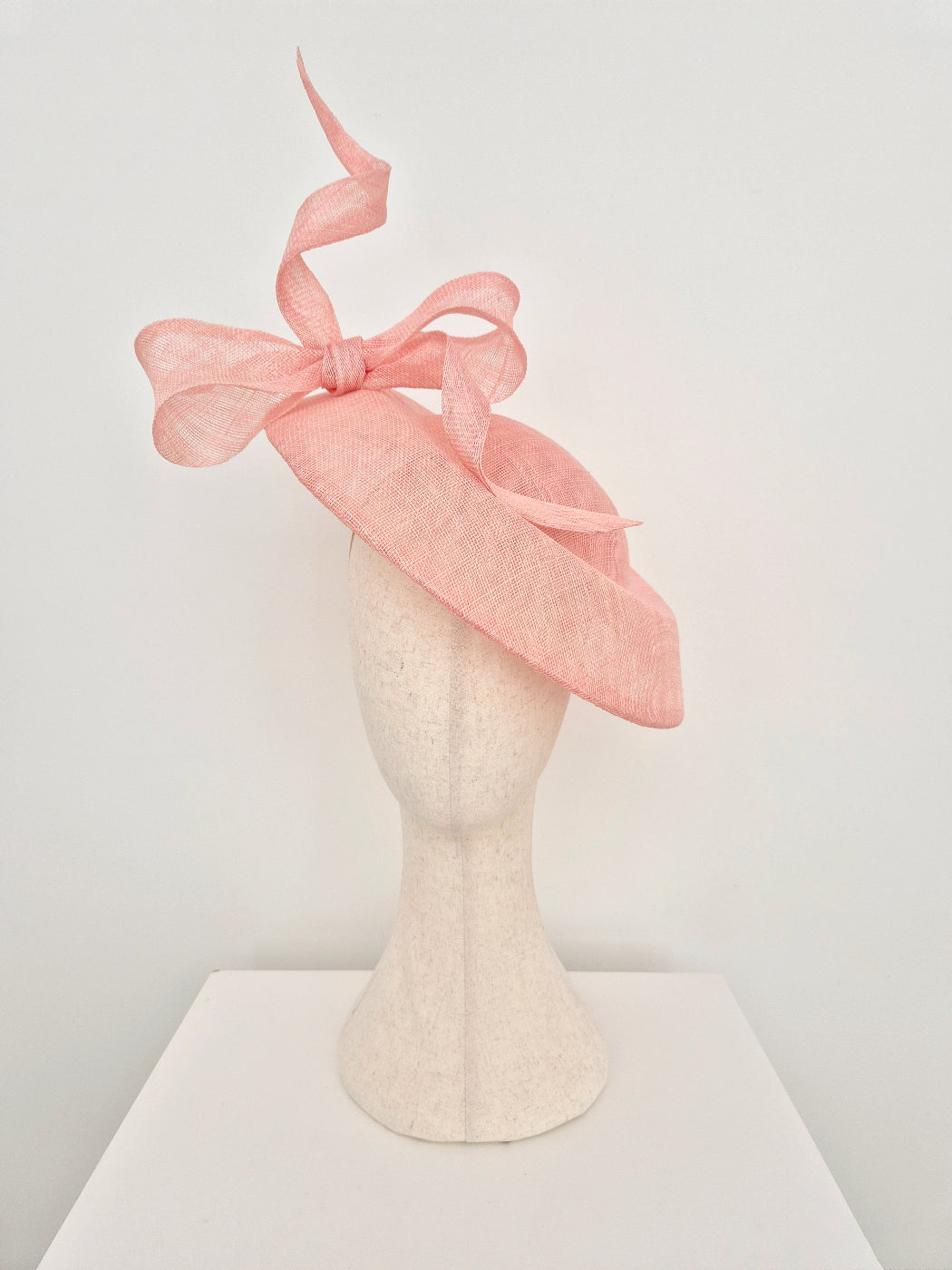 A light pink fascinator hat with a large bow and flowing ribbon, displayed on a textured cream mannequin head against a plain white background.