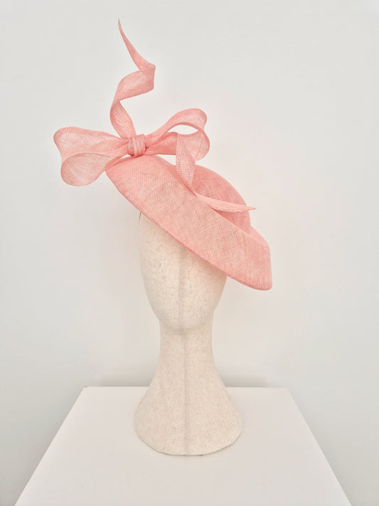 A light pink fascinator hat with a large bow and flowing ribbon, displayed on a textured cream mannequin head against a plain white background.
