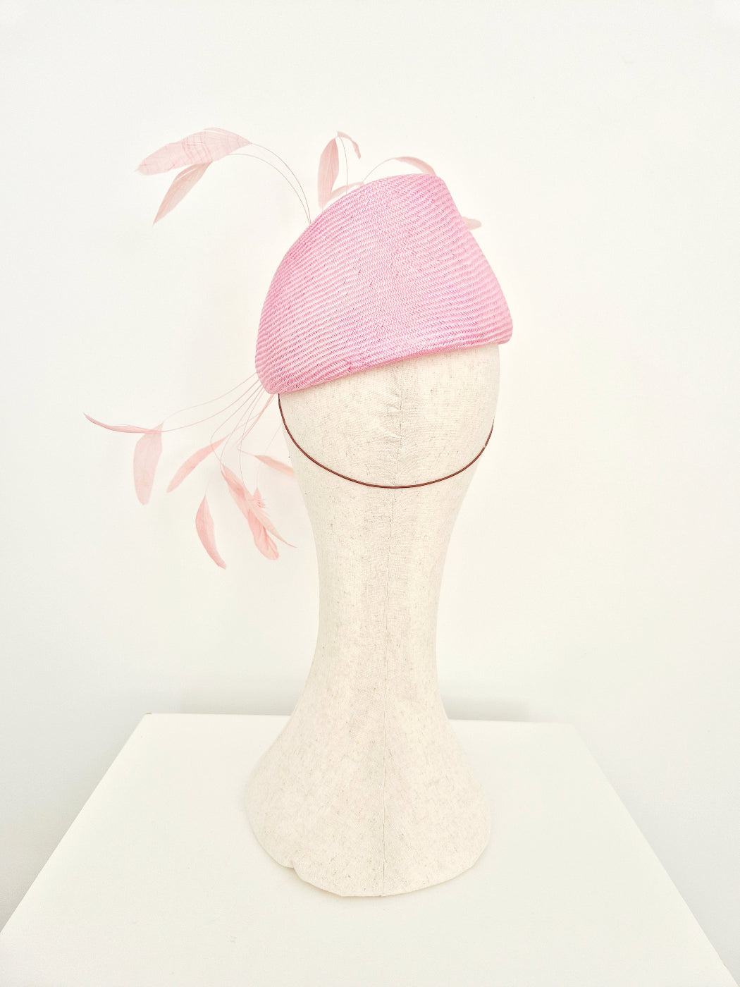 A light pink woven fascinator hat with delicate pale pink feather accents, displayed on a cream-colored felt mannequin head against a clean white background.back view