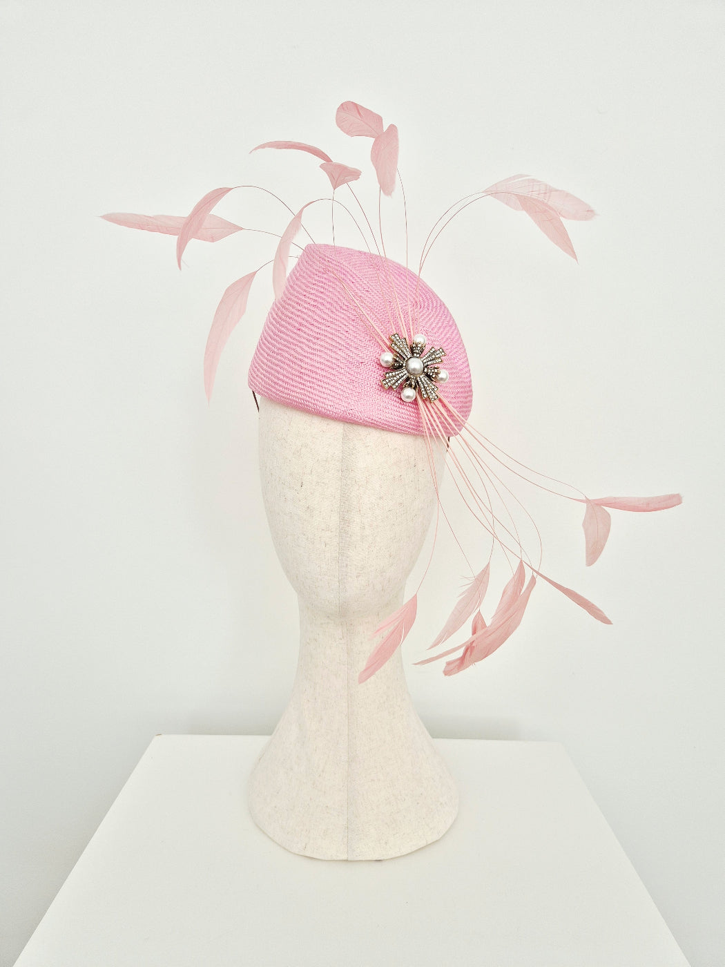 A light pink structured fascinator hat displayed on a cream-colored mannequin head. The hat features delicate pale pink feather accents and a decorative silver-toned floral embellishment with white pearls.