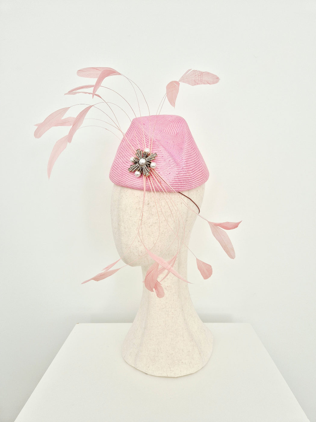 A pink fascinator hat with delicate feather accents and a jeweled center, displayed on a textured cream mannequin head against a clean white background. side view
