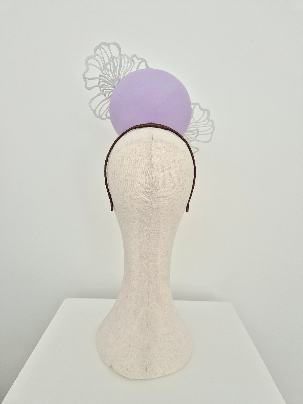 A lilac percher headband fascinator displayed on a cream-colored fabric mannequin head, set against a plain white wall back view