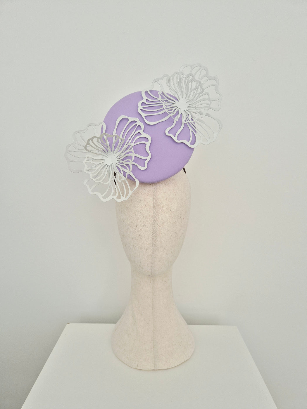 A lilac percher fascinator  with white laser-cut floral accents, displayed on a cream felt mannequin head against a light gray background.