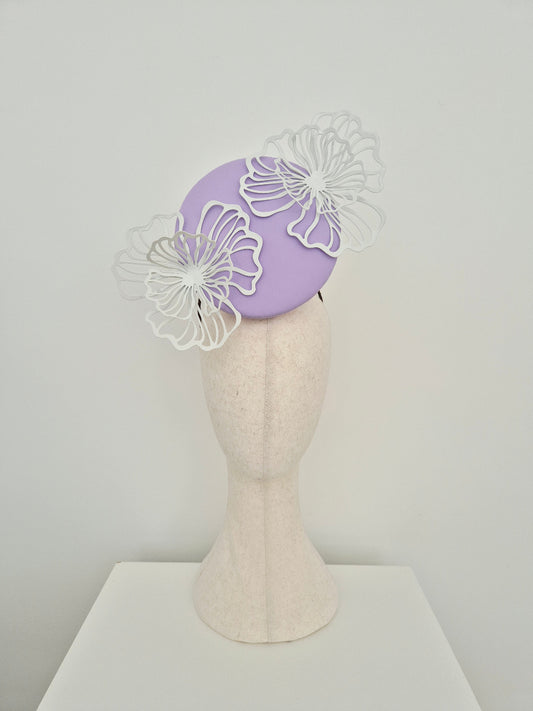 A lilac percher fascinator  with white laser-cut floral accents, displayed on a cream felt mannequin head against a light gray background.