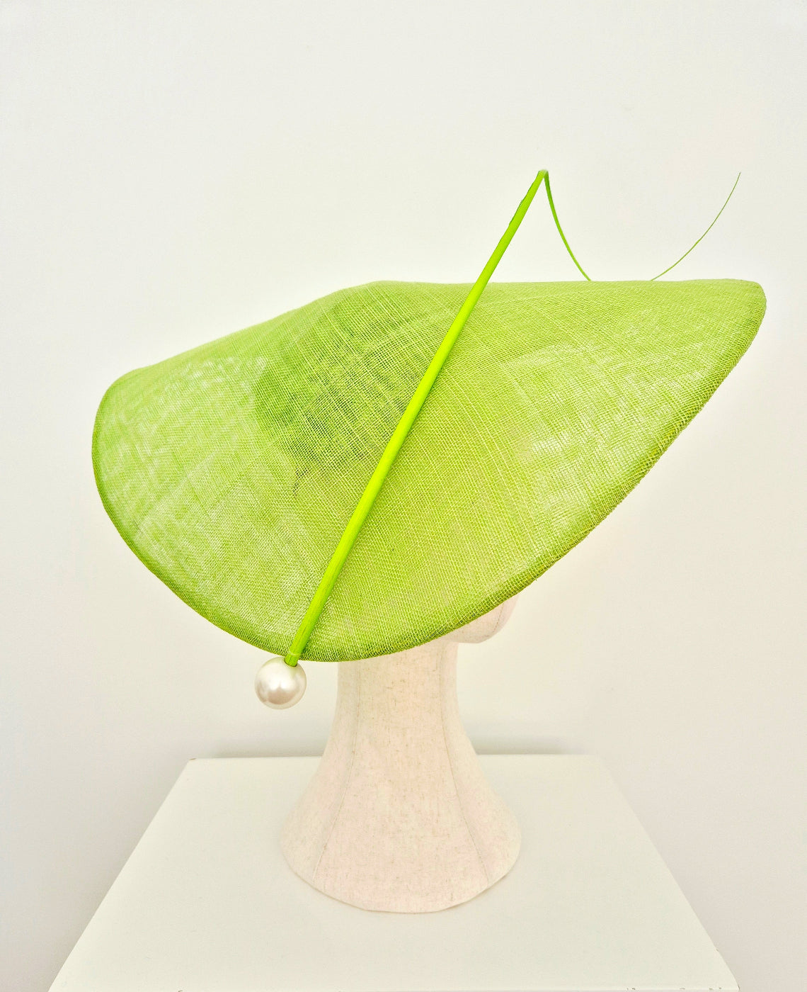 A bright lime green wide-brimmed fascinator hat with a long thin green feather accent and a  pearl bead, displayed on a light pink mannequin head against a white background. side view