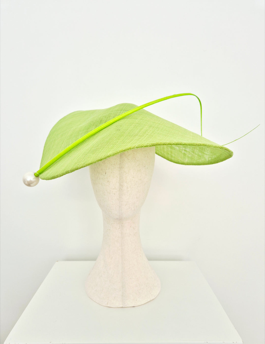 A light green fascinator hat with a curved brim, a thin matching green feather accent, and a white pearl bead on the side, displayed on a cream-colored mannequin head against a white background.straight on
