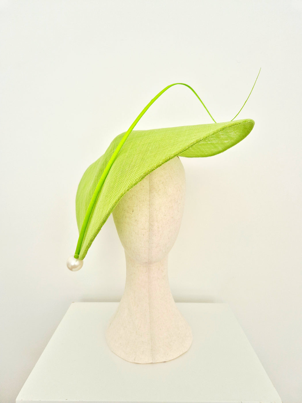 A bright lime green fascinator hat with a long, thin upward-curving stem and a small pearl bead accent, displayed on a textured off-white mannequin head against a clean white background.