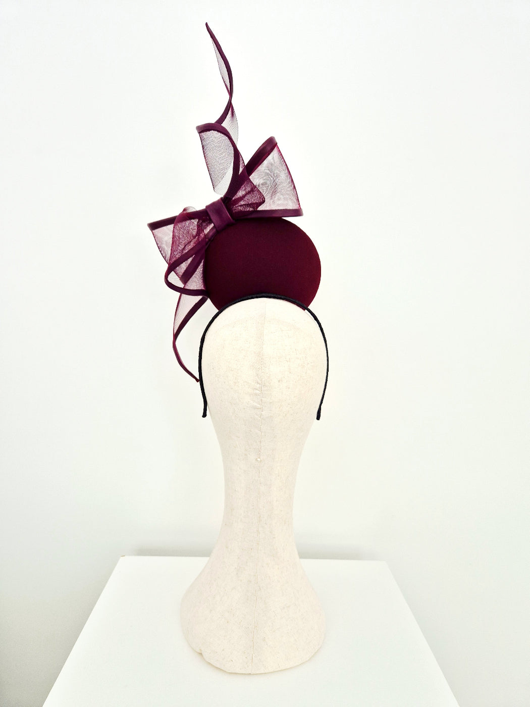 A fascinator hat displayed on a  mannequin head. The hat features a deep burgundy felt base with a matching burgundy sheer mesh bow and a tall, thin burgundy feather accent, attached to a thin black headband.Back view