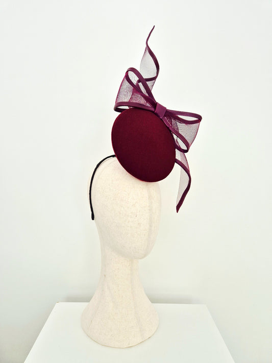 A deep burgundy felt fascinator hat with a matching sheer mesh bow and a black headband, displayed on a cream-colored mannequin head against a white background.side view