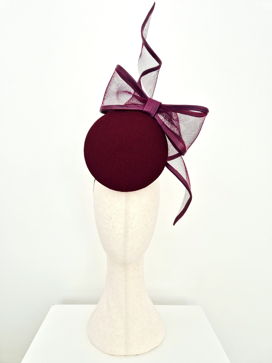 A dark burgundy felt fascinator hat with a matching sheer mesh bow, displayed on a cream-colored mannequin head against a plain white background.