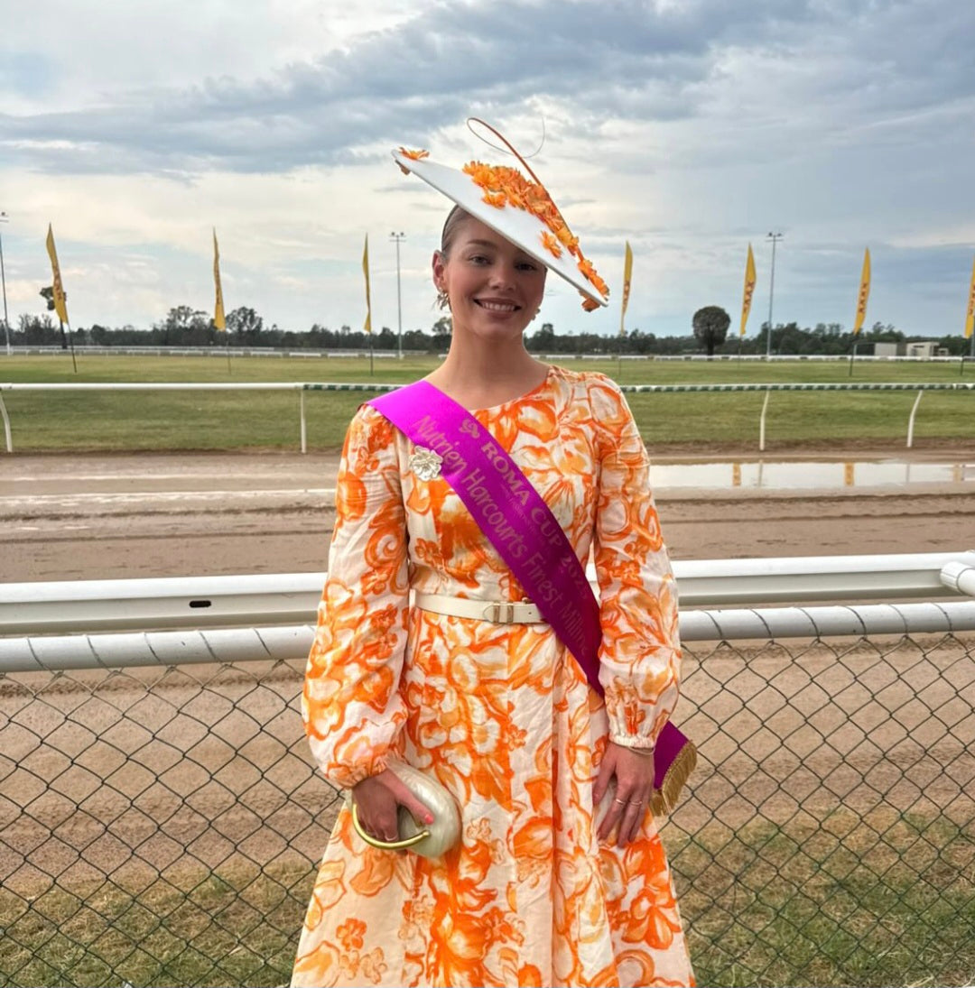 model wearing ivory and orange hat headpiece with a matching coloured dress at a race track