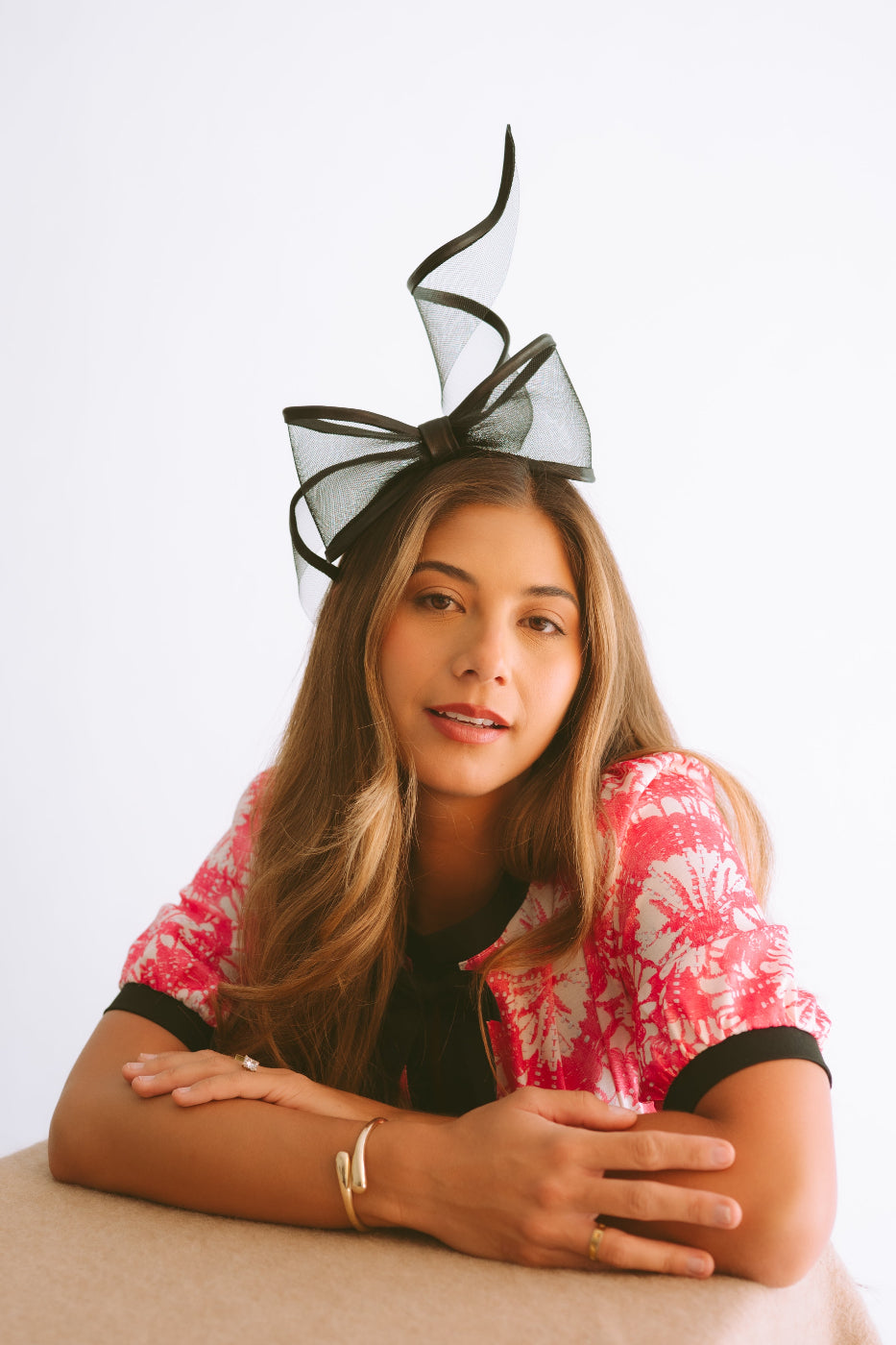 A woman with long light brown hair wearing a  black bow fascinator  and a pink and white patterned top, leaning on a beige table  against a white studio background.