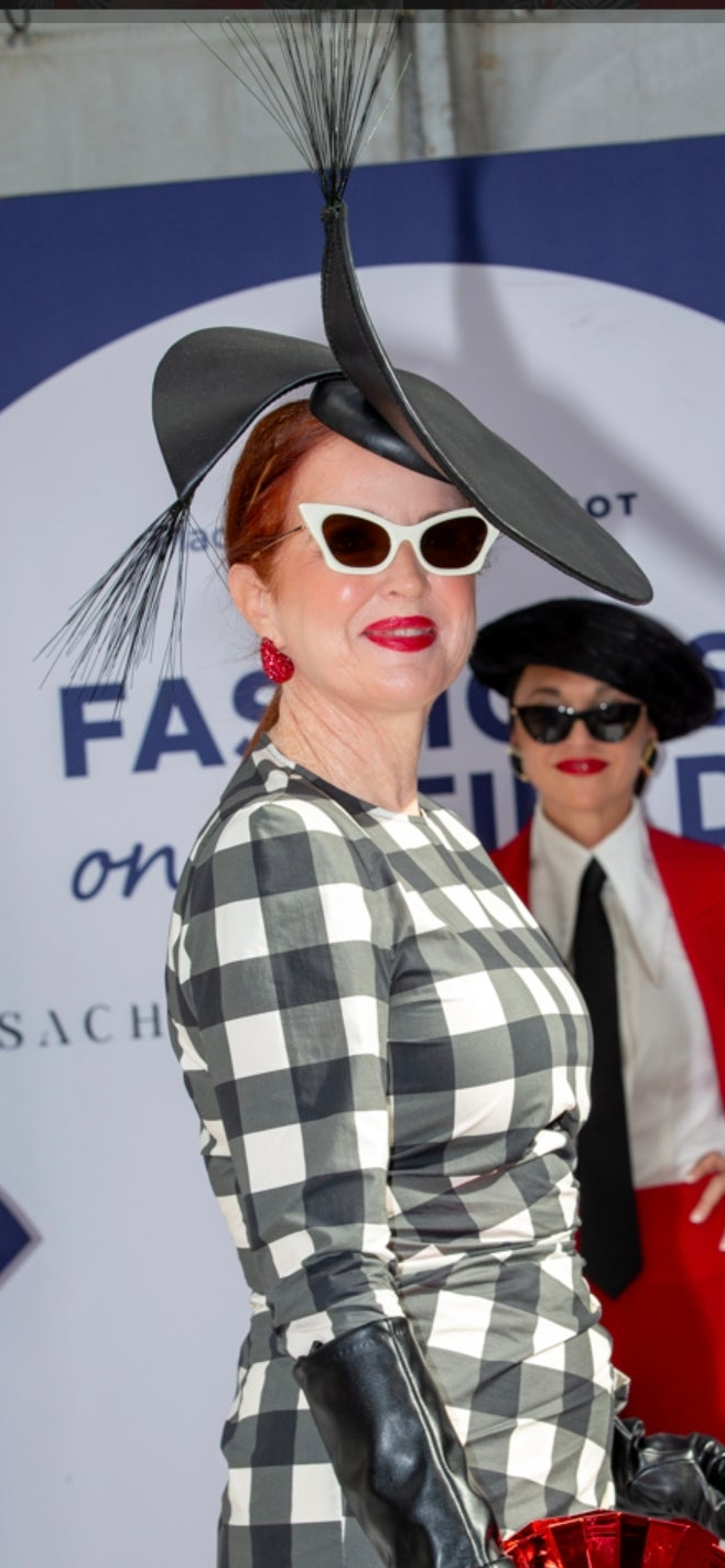 Person wearing a checkered outfit and large black hat with a feather, standing in front of a branded backdrop.