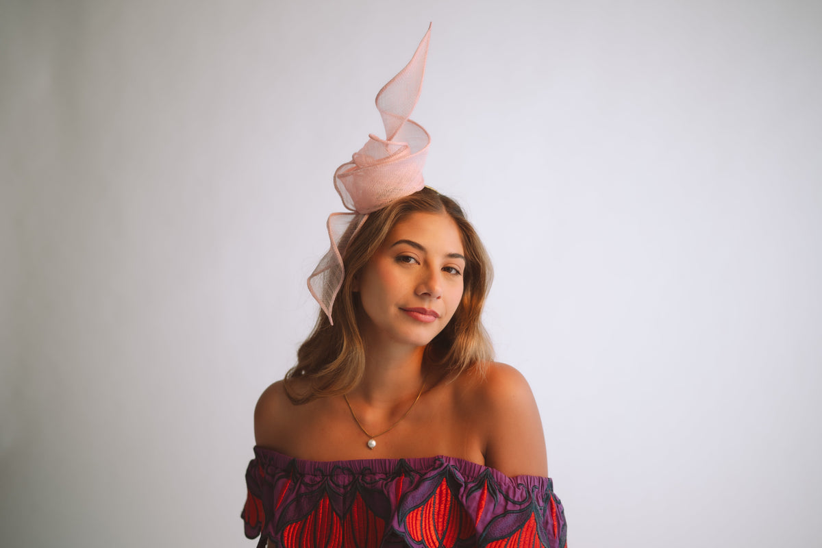 A woman wearing a light pink fascinator hat and a purple off-the-shoulder top with red patterns, photographed against a plain light gray background.