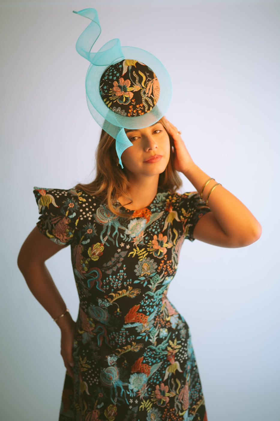A woman wearing a dark floral-patterned dress and a matching light blue fascinator hat, posing against a light gray studio background.