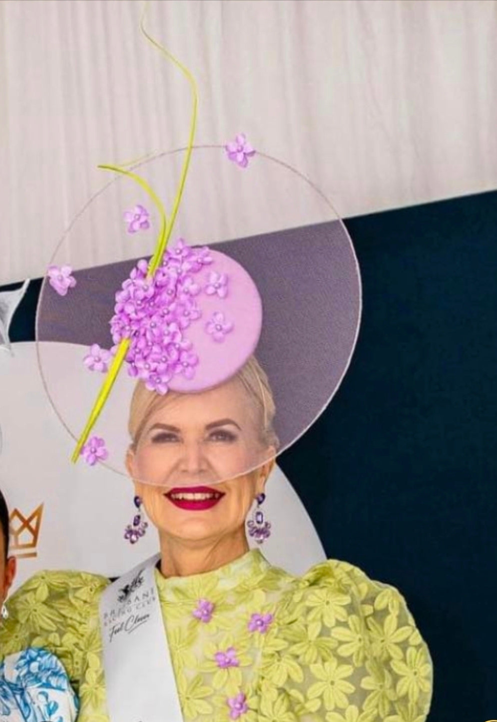 model wearing a decorative hat with flowers and a yellow outfit against a patterned background