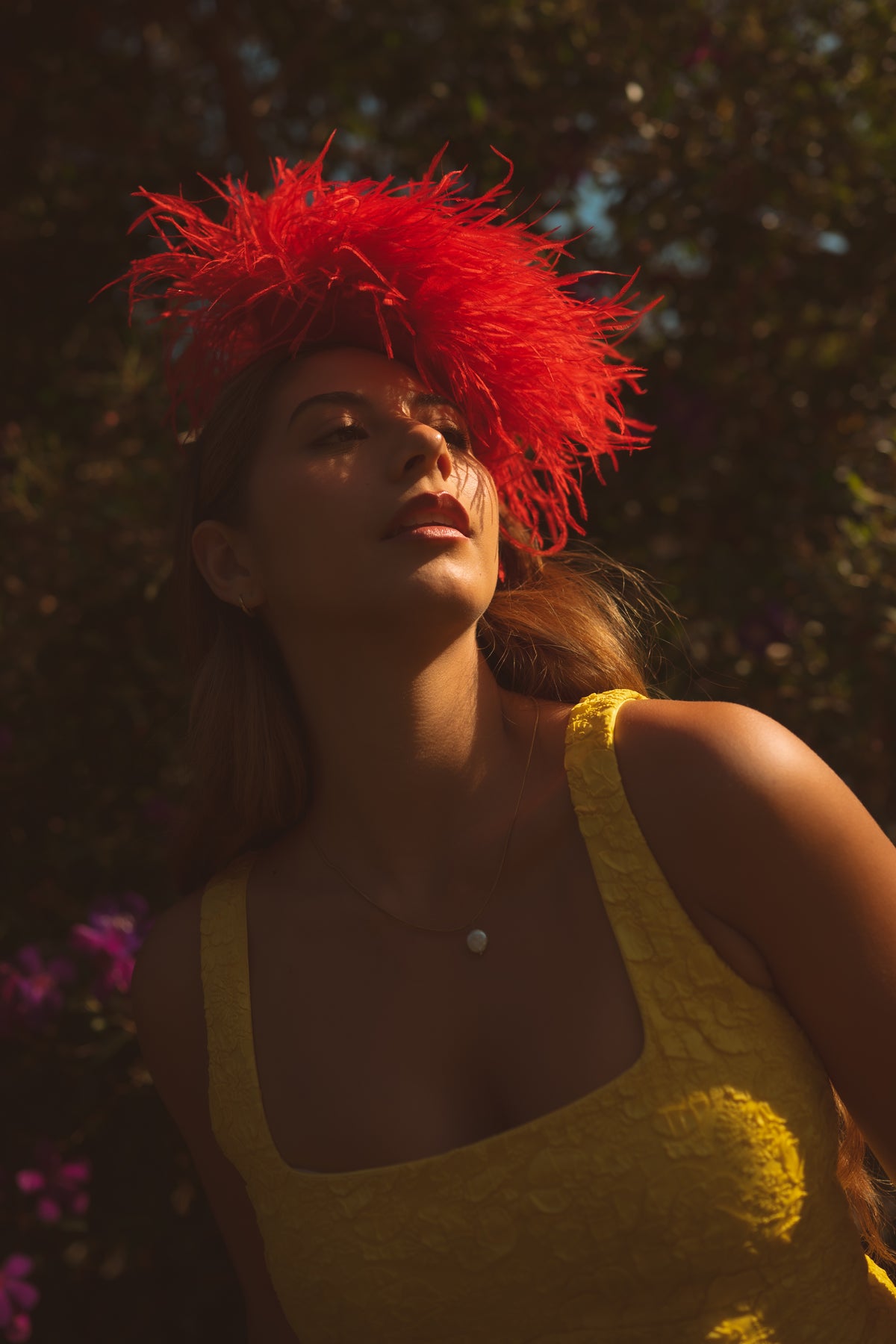 A woman outdoors wearing a bright red feather headpiece and a textured yellow top, with warm sunlight and dark foliage in the background.