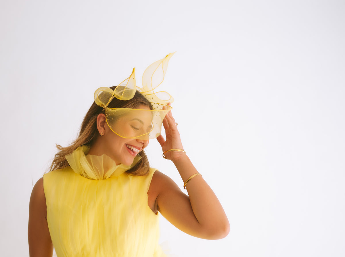 A woman wearing a bright yellow sleeveless dress and a matching yellow fascinator headpiece, posing against a solid white studio background.