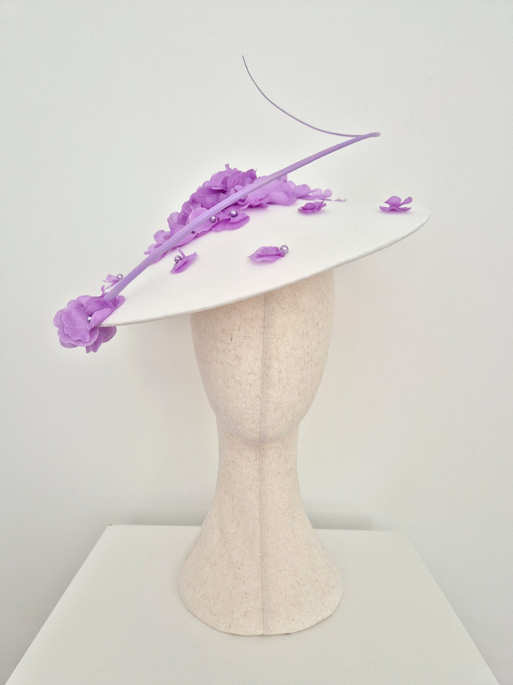 A white fascinator hat decorated with purple flowers and a long purple feather, displayed on a textured cream-colored mannequin head against a plain white background.