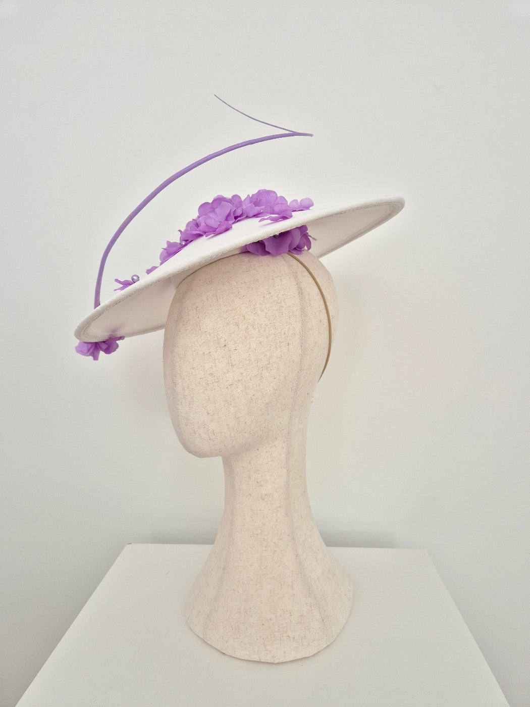 A white fascinator hat with purple floral accents and a curved purple feather plume, displayed on a beige felt mannequin head against a plain white background.underside view
