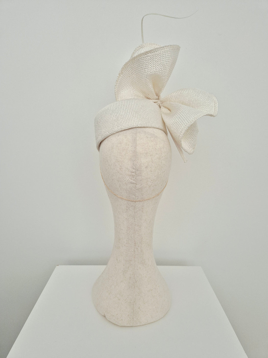 An off-white straw textured fascinator hat with a large bow and a thin curved quill accent, displayed on a matching off-white mannequin head stand against a light gray background. back view