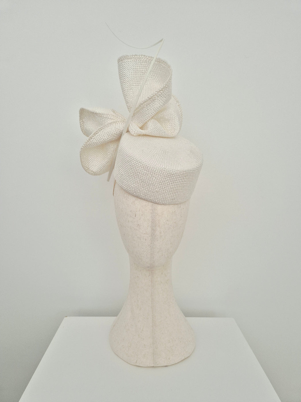 An off-white textured straw fascinator  with a large bow and quill, displayed on a matching off-white mannequin head stand against a plain light gray background.