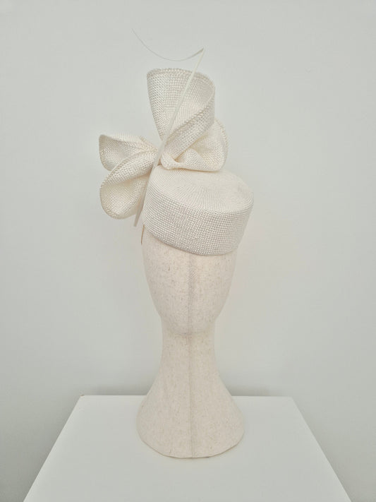 An off-white textured straw fascinator  with a large bow and quill, displayed on a matching off-white mannequin head stand against a plain light gray background.