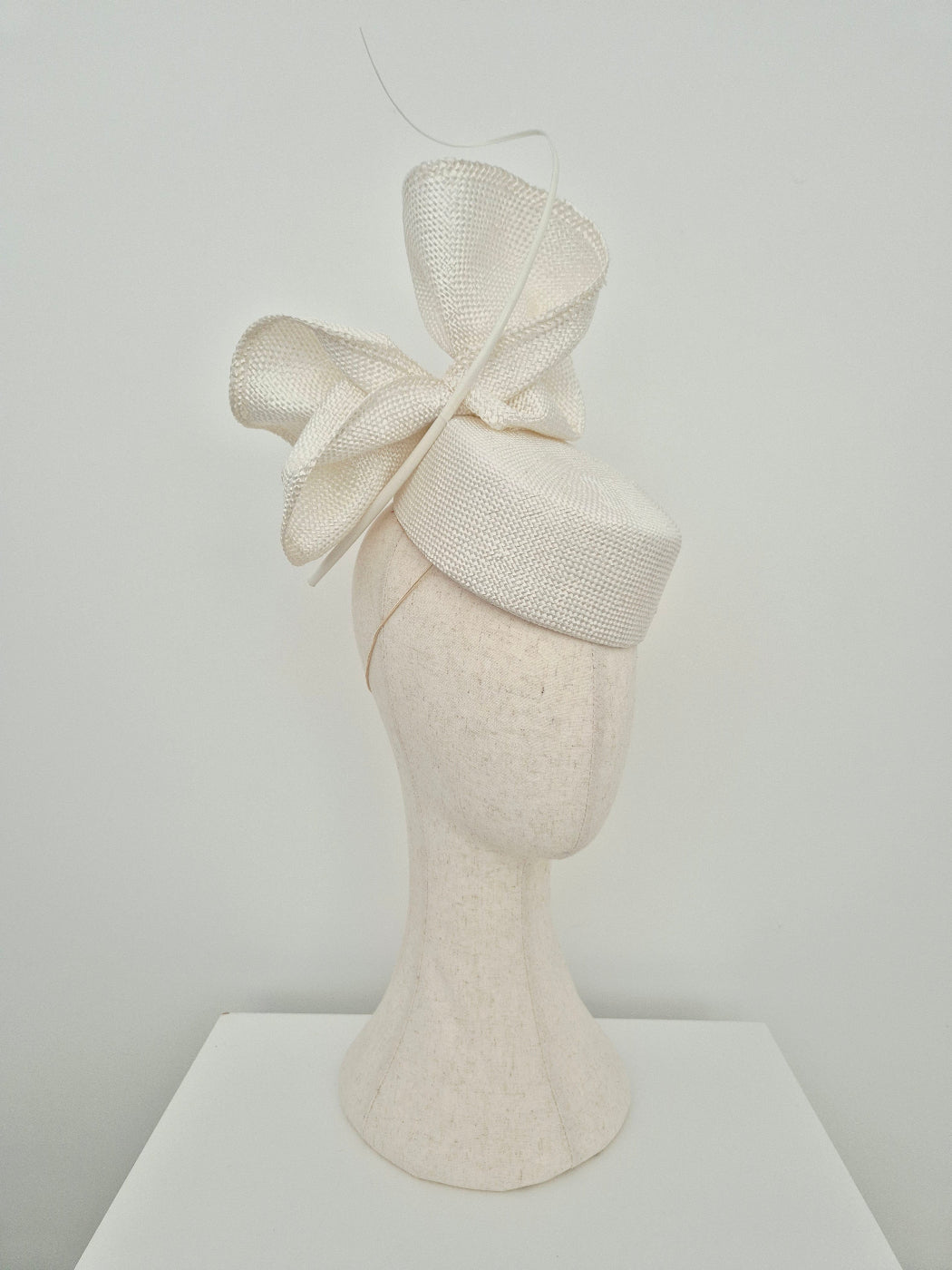 An off-white textured straw fascinator hat with a large bow, displayed on a matching off-white felt mannequin head against a plain light gray background. side view