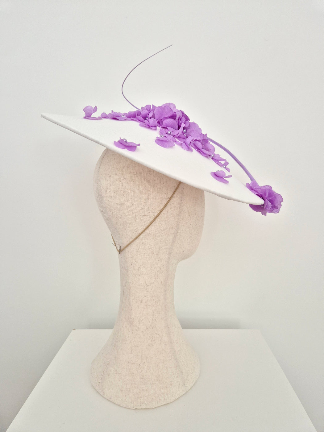 A white fascinator hat with purple floral embellishments and a long thin feather, displayed on a beige mannequin head against a plain white background.side view 2