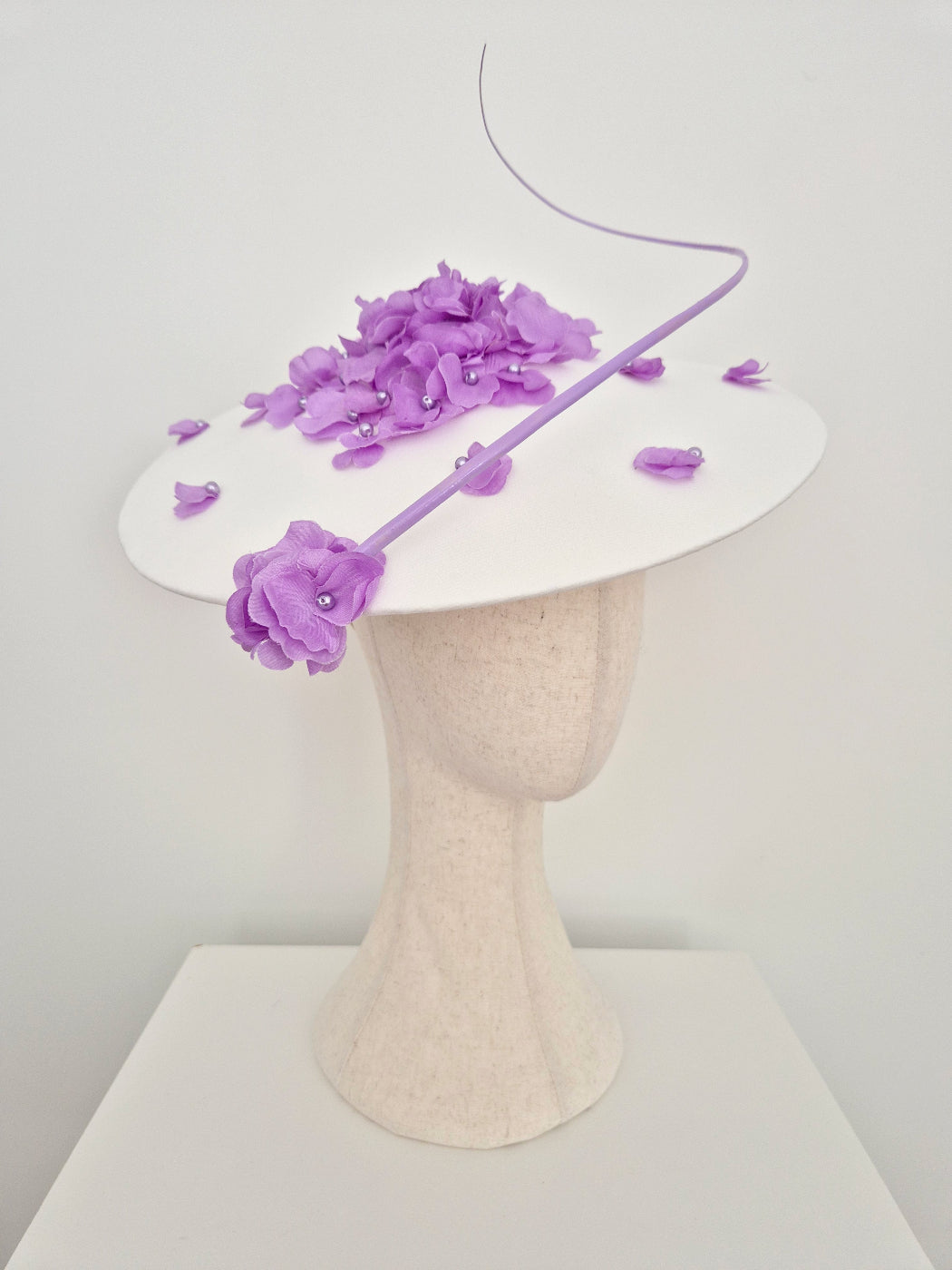 A white fascinator hat displayed on a textured cream mannequin head, decorated with purple fabric flowers, petals, and a thin purple wire accent against a plain white background.side front view