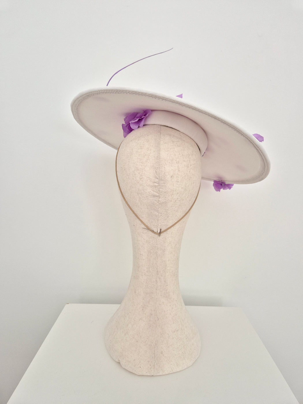 A off white fascinator hat with a wide brim and small purple floral accents, displayed on a cream-colored felt mannequin head against a plain white background.back view