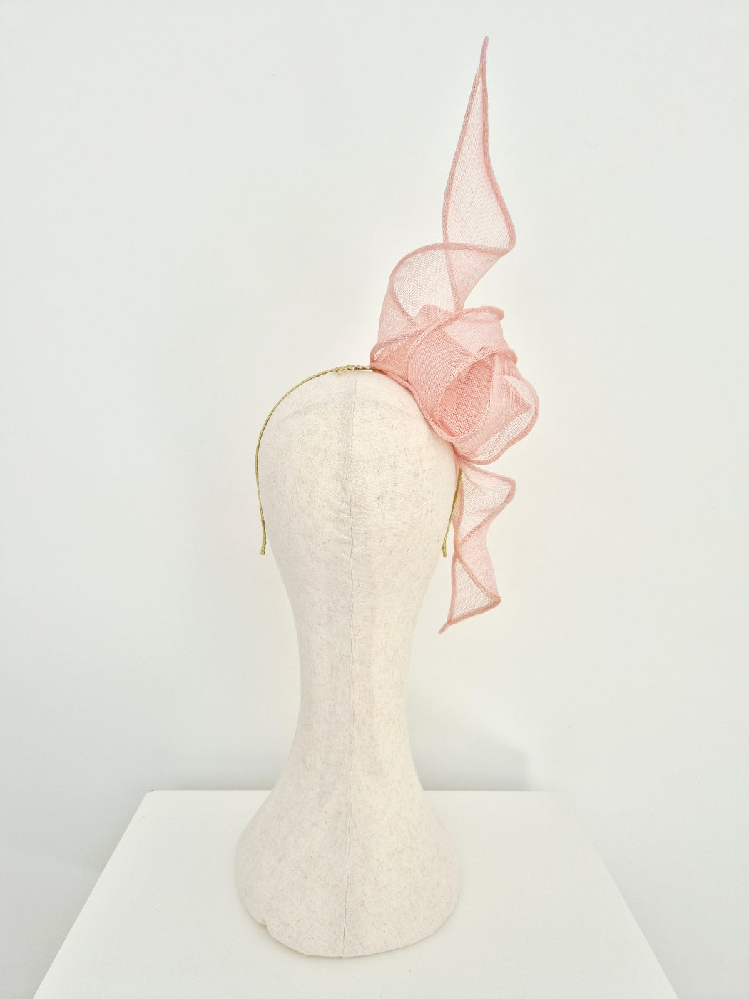 A pale pink fascinator hat with a sheer straw knott and tails on a  mannequin head against a white background. back view