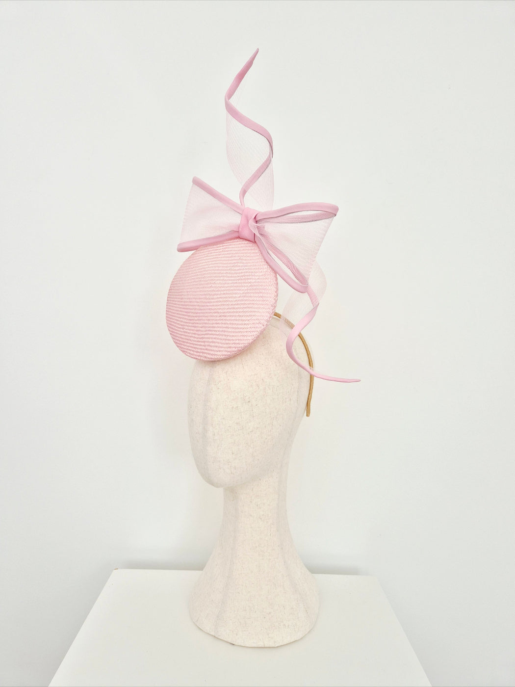 A light pink straw percher fascinator headpiece with a large bow on a mannequin head against a white background. side view
