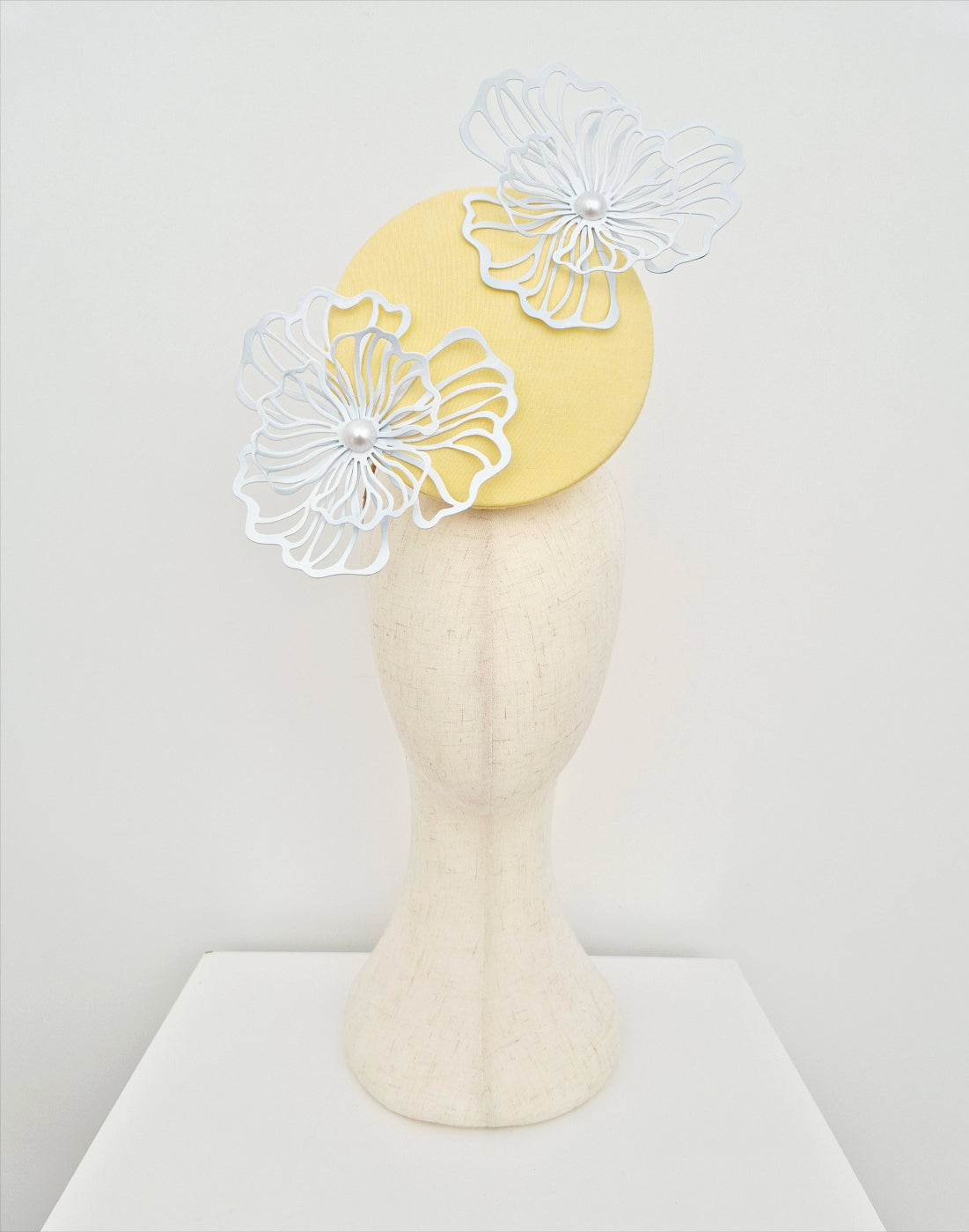 Yellow fascinator with white floral decorations on a mannequin head against a white background
