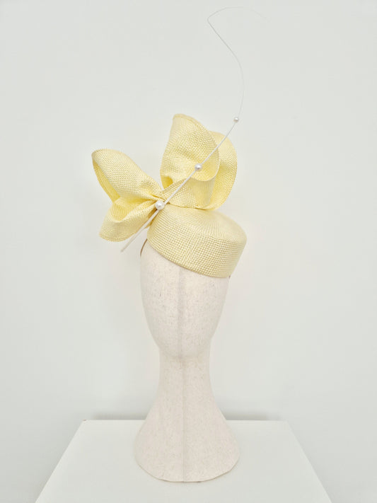 A pale yellow straw pillbox fascinator hat with a large bow and quill accent, displayed on a textured off-white mannequin head against a clean white background. tilted