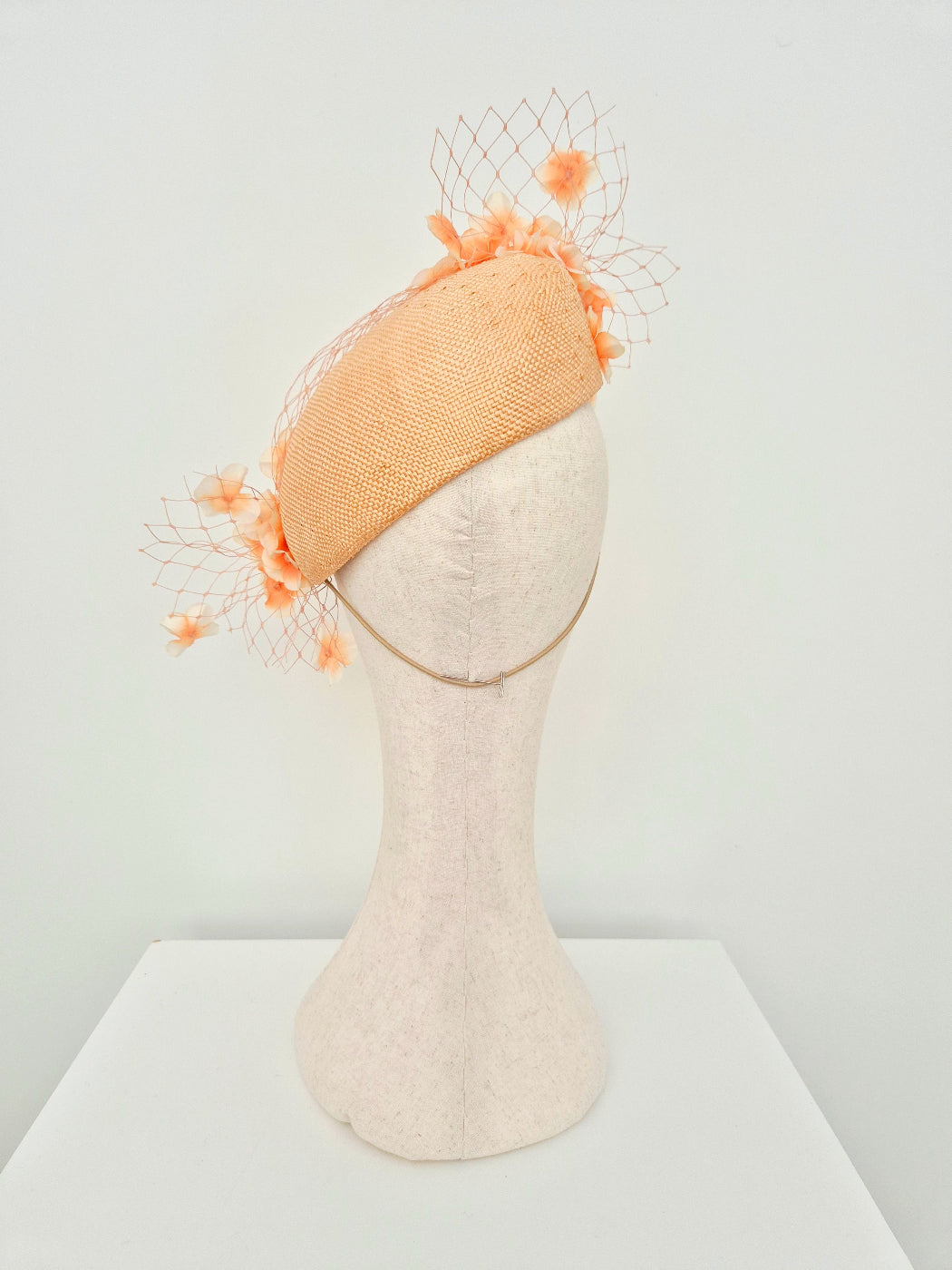 A peach-colored straw fascinator hat with matching orange floral accents and a delicate birdcage net, displayed on a cream-colored mannequin head against a white background.back view