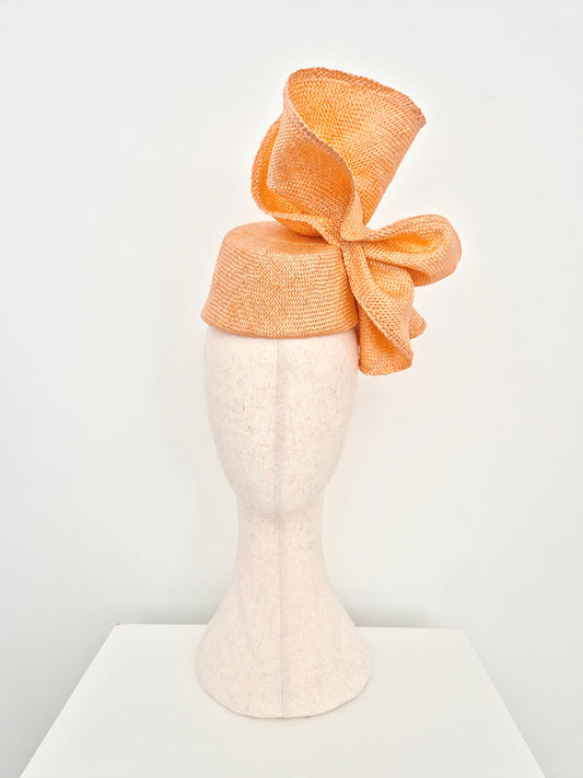 An peach straw fascinator hat with a large bow, displayed on a textured off-white mannequin head against a clean white background.front on