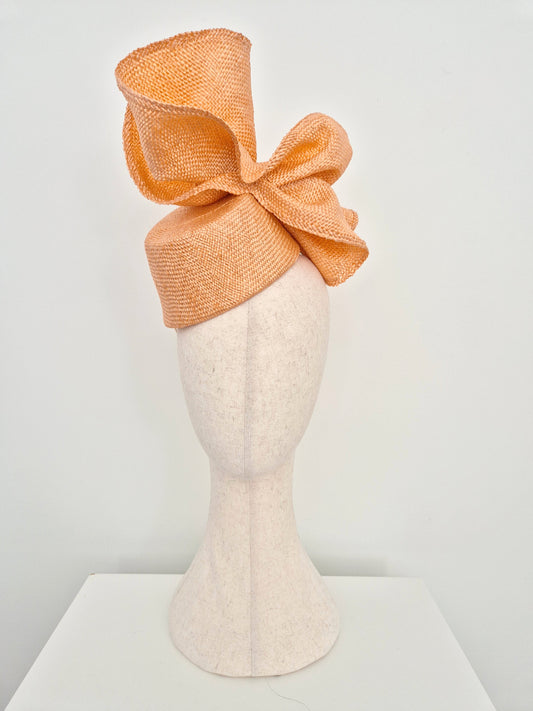 a peach straw fascinator hat with a large bow, displayed on a cream-colored felt mannequin head against a white background.tilted