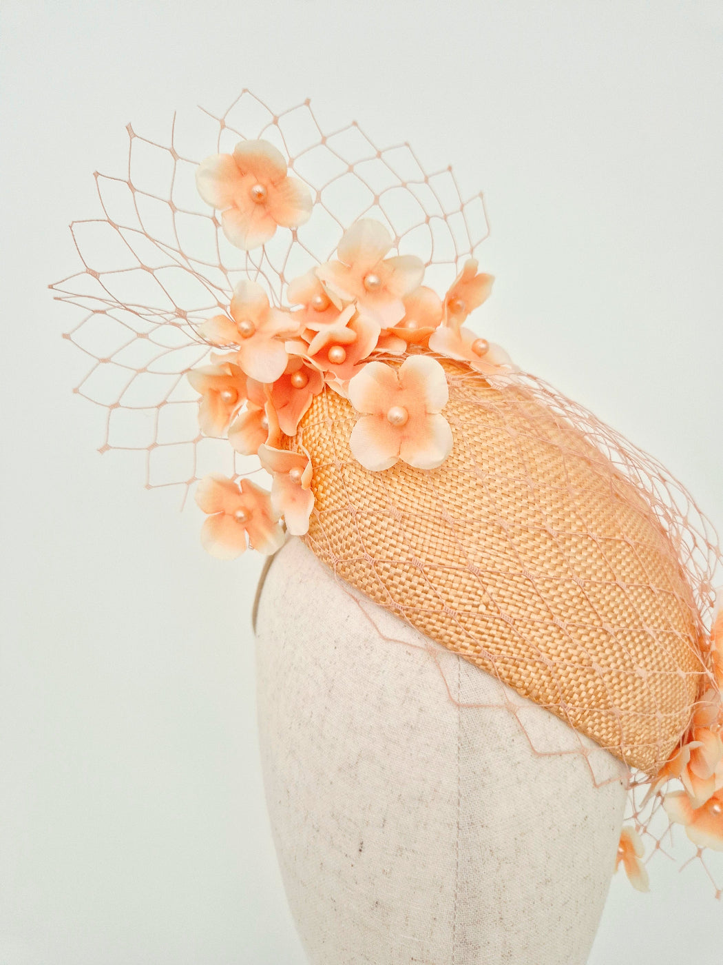 A peach-colored woven fascinator hat with delicate floral embellishments and a matching net veil, displayed on a textured mannequin head against a light gray background.close up
