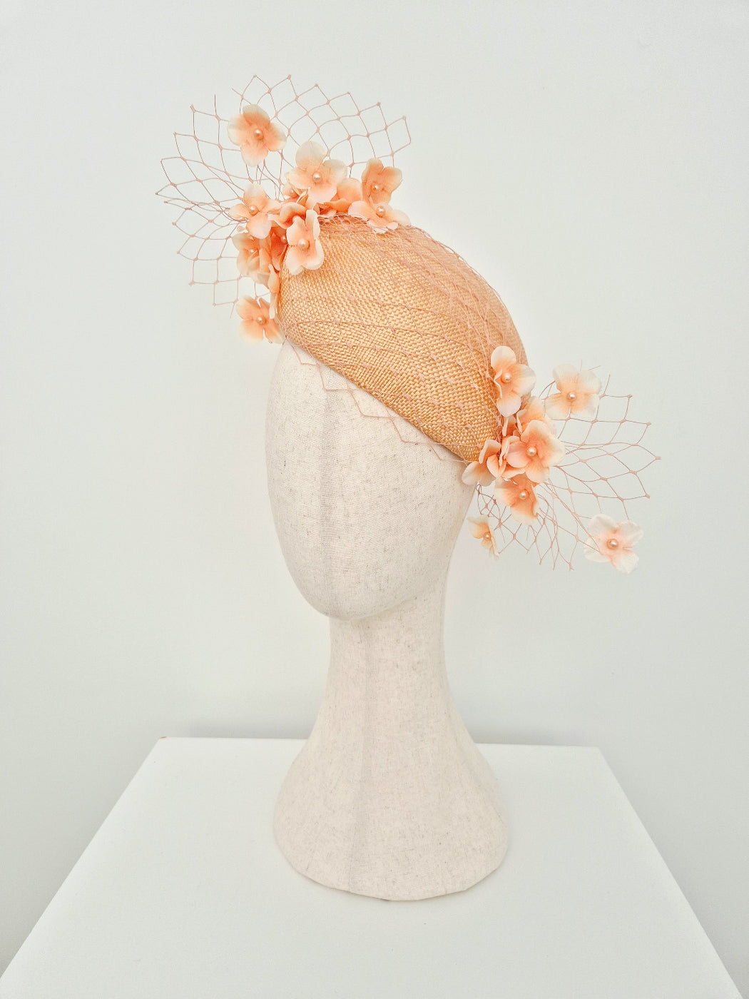 A peach-colored fascinator with floral embellishments and a delicate netting veil, displayed on a textured off-white mannequin head against a clean white background. side view