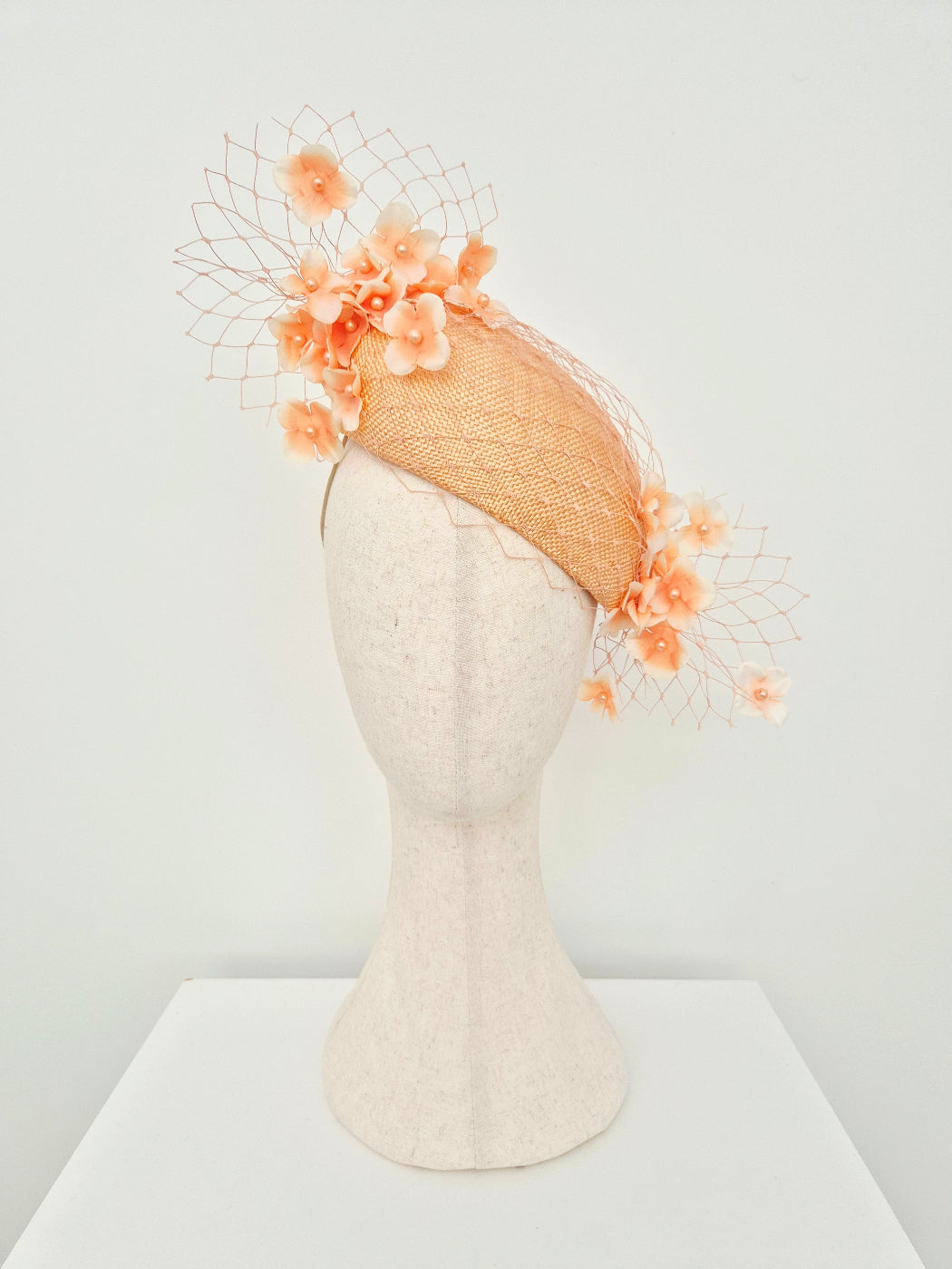A peach colored straw fascinator hat with attached orange artificial flowers and a fine mesh veil, displayed on a textured off-white mannequin head against a plain white background.