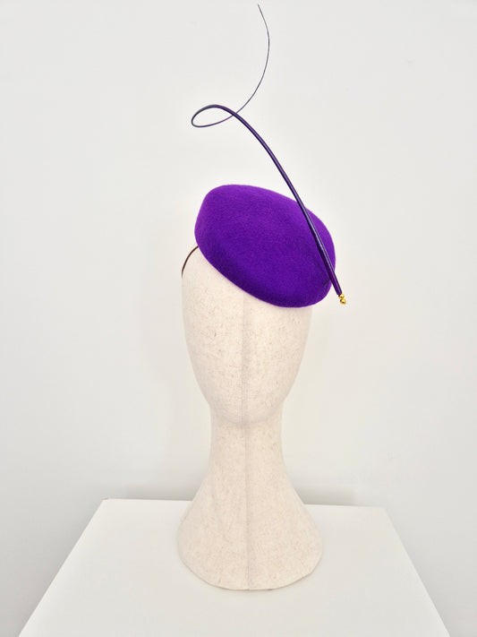 A purple felt fascinator with a long, sweeping quill and a tiny gold tip, displayed on a cream-colored felt mannequin head against a clean white background.
