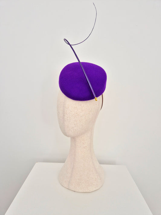 A  purple felt fascinator hat with a single sweeping feather plume, displayed on a textured cream-colored mannequin head against a clean white background.side view