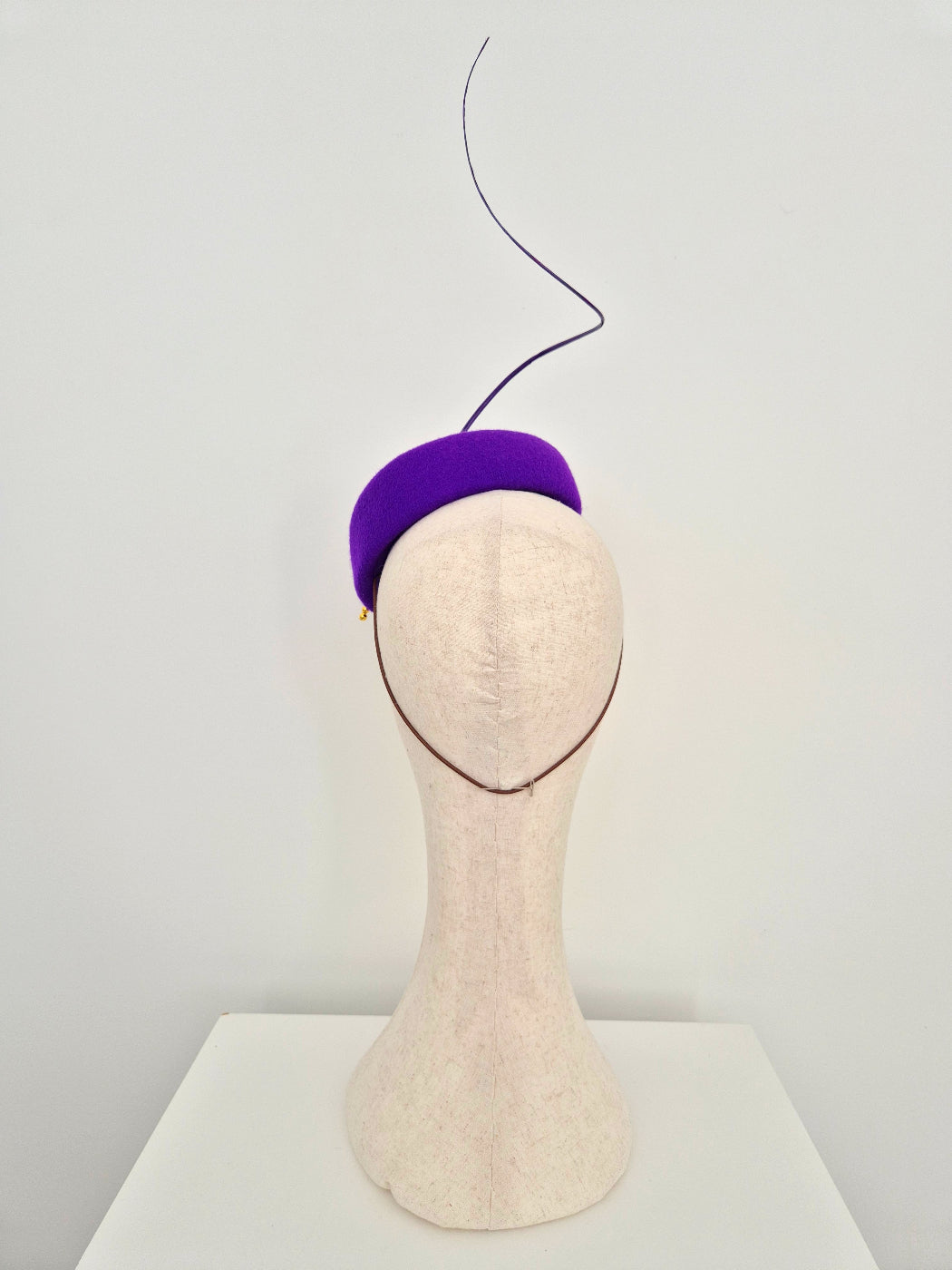 A purple felt fascinator  with a quill displayed on a beige mannequin head against a white background. back view